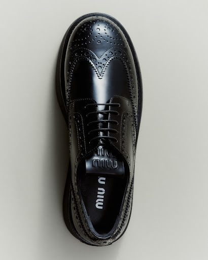 Studio shot of a black, patent-leather wingtip-style lace-up shoe with a thick lug sole from Miu Miu, positioned against a neutral backdrop.