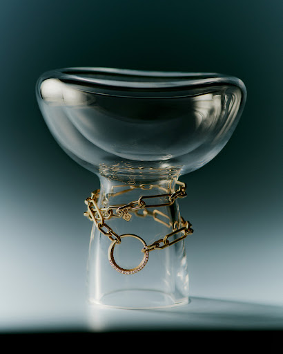 Clear glass bowl and short glass stand with gold chain necklace around the stand on a graded teal to light blue background.