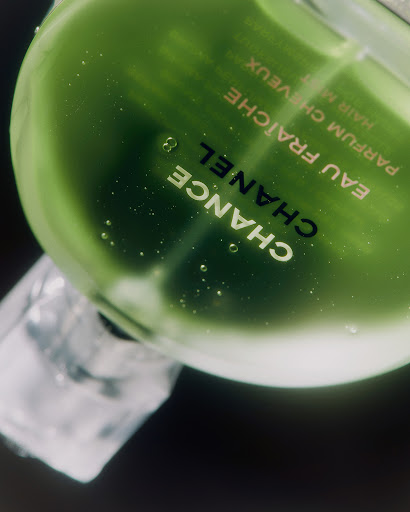 Close-up of a green Chanel Chance Eau Fraiche hair mist bottle with visible liquid and product branding, on a dark surface.
