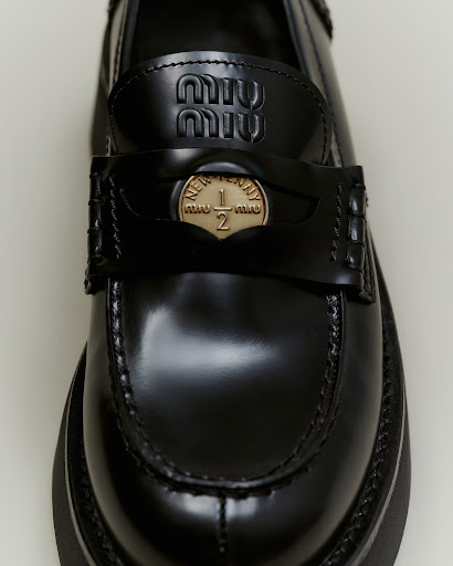 Close-up of a black leather Miu Miu loafer with a gold coin detail, showcasing the brand's logo and stylish design on a neutral background.