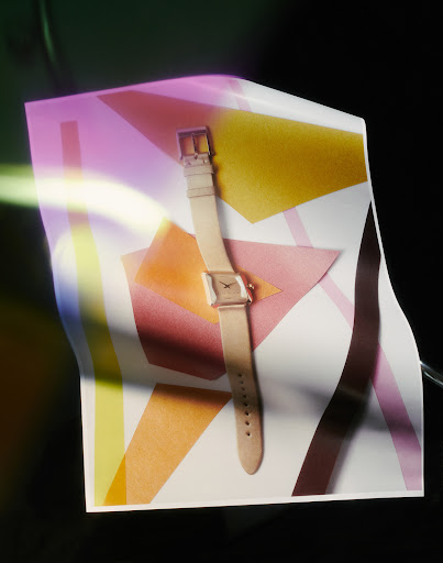 Elegant wrist watch with a neutral band and square face on a geometric white and pastel patterned surface in a studio setting.