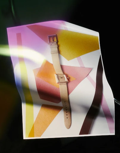 Elegant wrist watch with a neutral band and square face on a geometric white and pastel patterned surface in a studio setting.