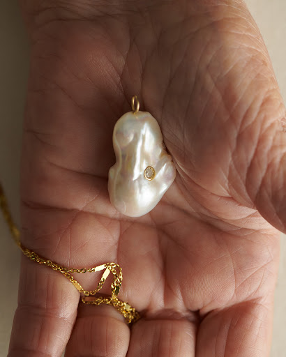 Close-up of a person's hand holding a baroque pearl pendant with a gold chain, showcasing the details of the jewelry.