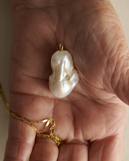 Close-up of a person's hand holding a baroque pearl pendant with a gold chain, showcasing the details of the jewelry.