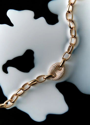 Elegant gold chain necklace with a diamond-encrusted clasp, set against a contrasting black background partially covered in white liquid.