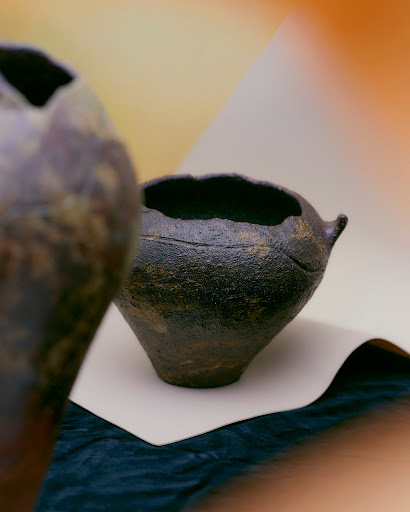 Two brown handmade ceramic pots sit on a white cloth against a blurred gradient background in shades of brown and orange.