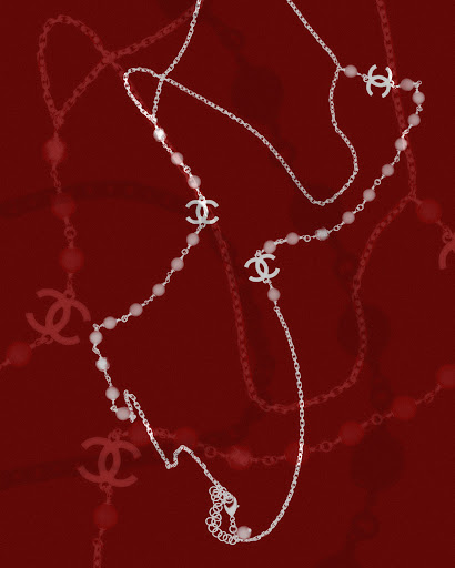 A delicate Chanel long necklace features a silver chain, pearl beads, and Chanel logo charms, set against a deep red background.