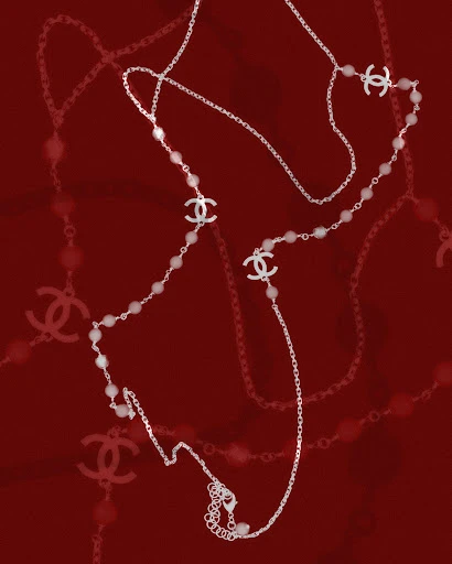A delicate Chanel long necklace features a silver chain, pearl beads, and Chanel logo charms, set against a deep red background.