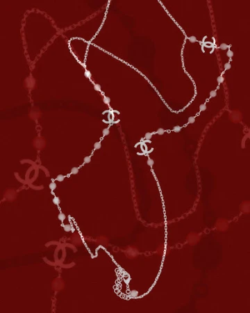 A delicate Chanel long necklace features a silver chain, pearl beads, and Chanel logo charms, set against a deep red background.