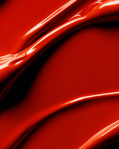 A close-up shot of thick, glossy red liquid, possibly paint or cosmetic product, flowing and rippling across the frame.