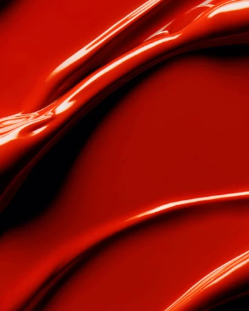 A close-up shot of thick, glossy red liquid, possibly paint or cosmetic product, flowing and rippling across the frame.