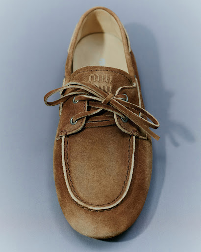 Close-up studio shot of a tan suede boat shoe with white trim and laces against a gray background, featuring the 'Miu Miu' logo.