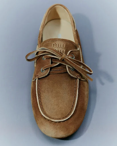 Close-up studio shot of a tan suede boat shoe with white trim and laces against a gray background, featuring the 'Miu Miu' logo.