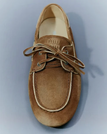 Close-up studio shot of a tan suede boat shoe with white trim and laces against a gray background, featuring the 'Miu Miu' logo.