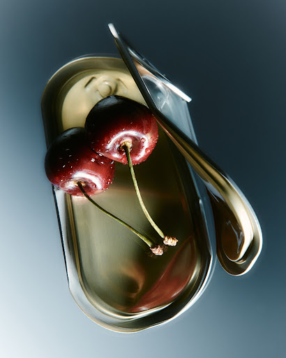 High-angle studio shot of two cherries with stems resting on an open, reflective tin can against a graduated blue backdrop.