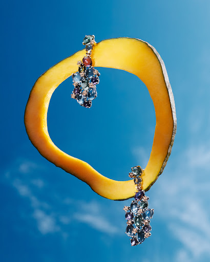 Elegant drop earrings featuring blue and purple gems dangle against a bright blue sky with a yellow squash rind accent.