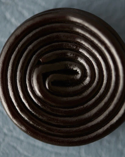 Close-up of a shiny, tightly-coiled spiral of black licorice candy on a textured light blue surface, showcasing its glossy finish.