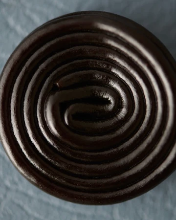 Close-up of a shiny, tightly-coiled spiral of black licorice candy on a textured light blue surface, showcasing its glossy finish.
