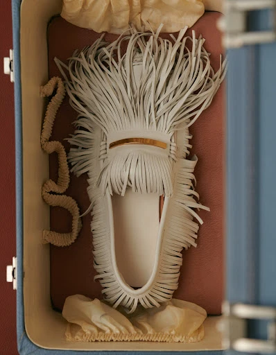 Close-up studio shot of a white, fringed, slip-on shoe inside of a brown, blue, and tan suitcase with cream-colored fabric. 