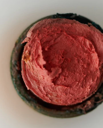 Overhead close-up of textured, red, spherical lip balm or rouge inside a green and black container, set against a white background.