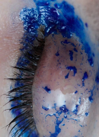 Extreme close-up of closed human eyelid covered in textured blue pigment and long dark eyelashes captured by Sara Zanoni