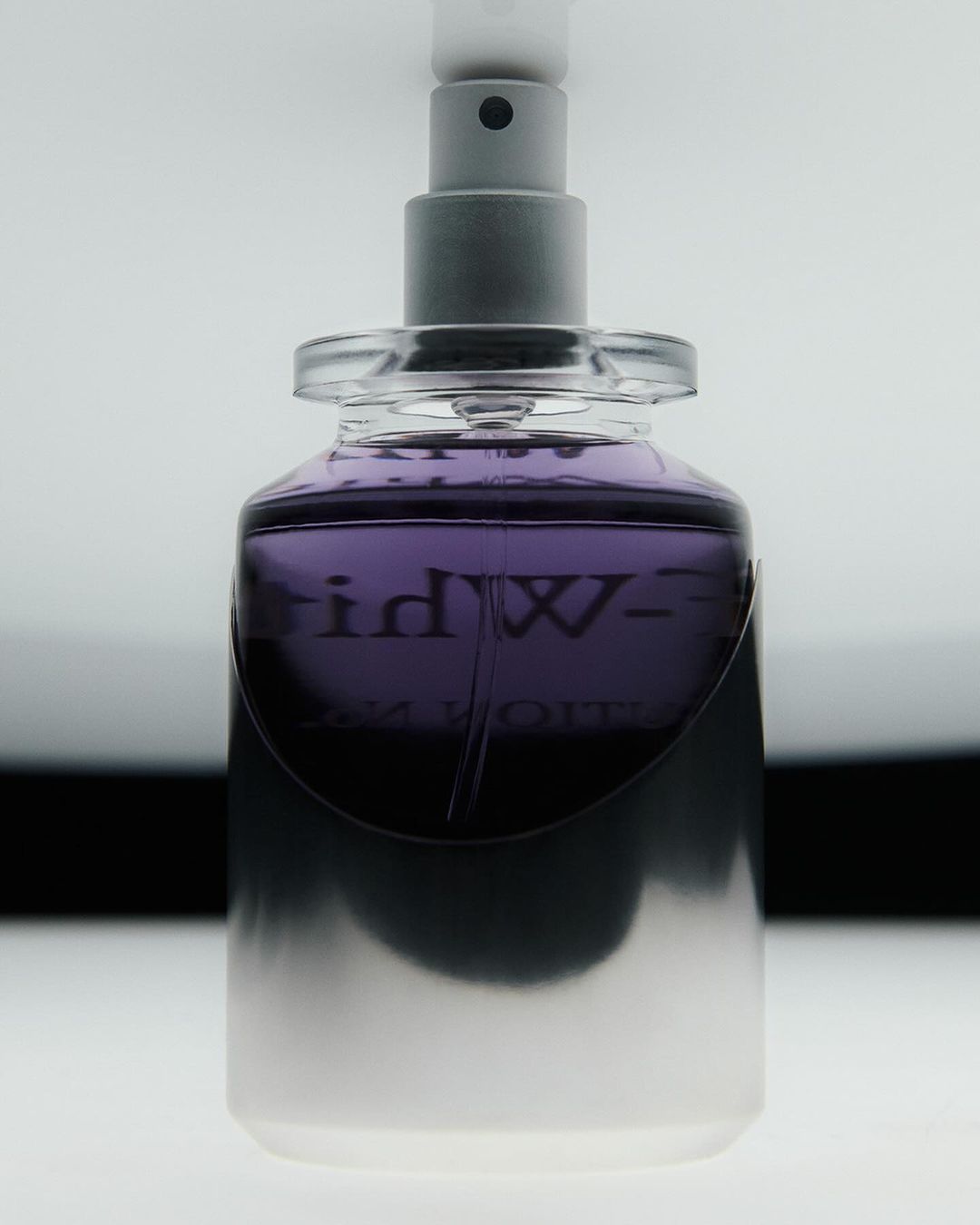 Close-up shot of a purple-tinted glass perfume bottle with a silver sprayer top against a gradient white to black background.