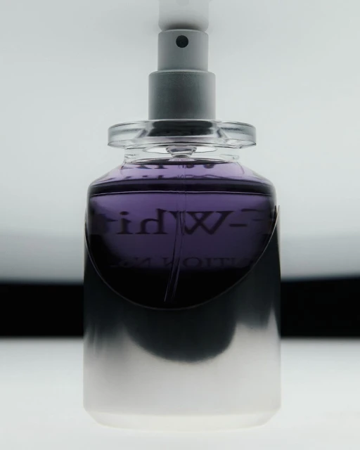Close-up shot of a purple-tinted glass perfume bottle with a silver sprayer top against a gradient white to black background.