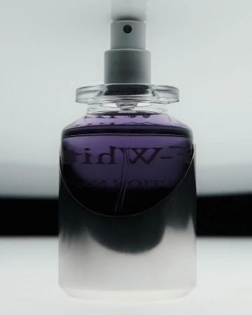 Close-up shot of a purple-tinted glass perfume bottle with a silver sprayer top against a gradient white to black background.