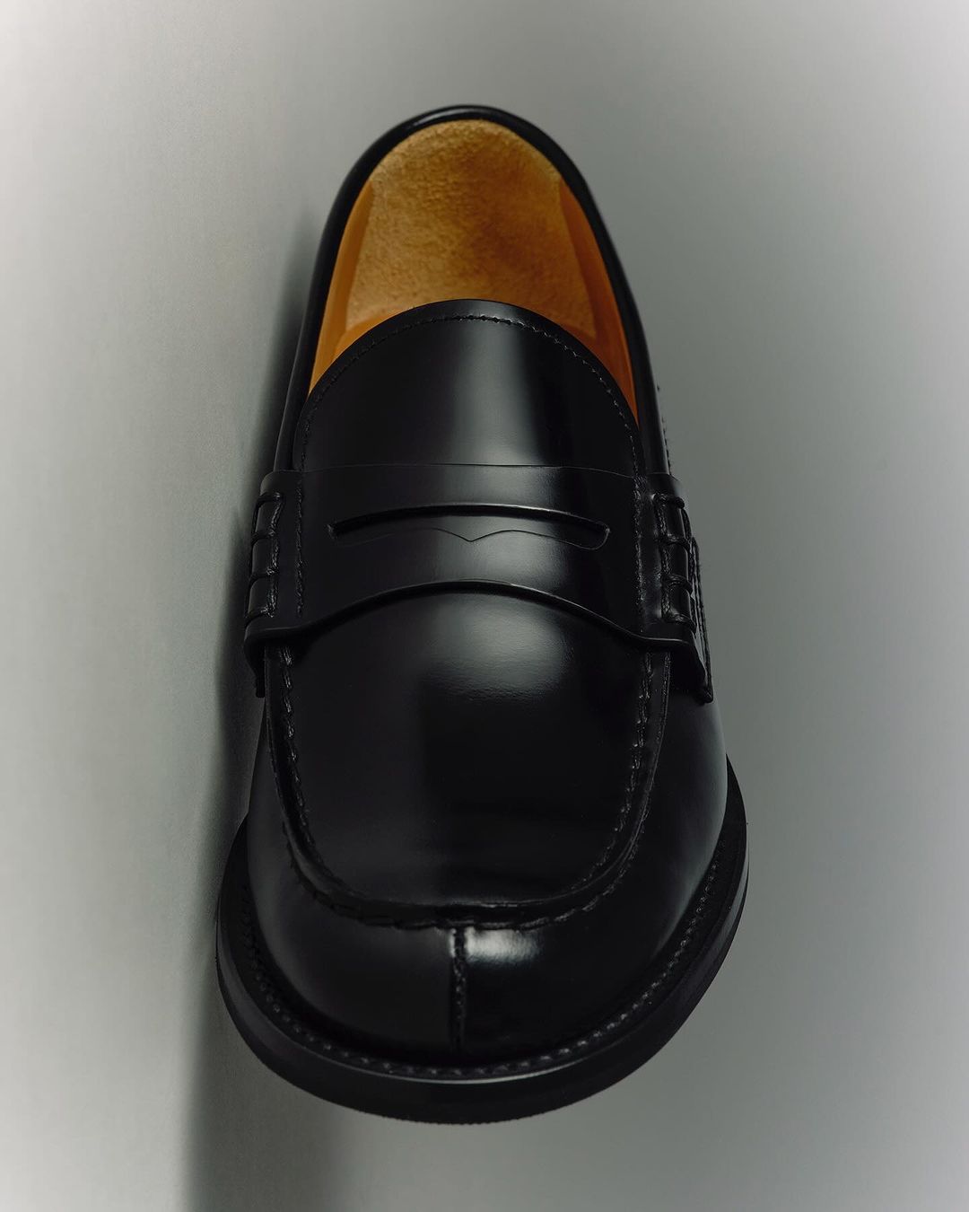 Closeup of a shiny black leather penny loafer shoe on a white background with smooth texture and light shadowing, showcasing its design.