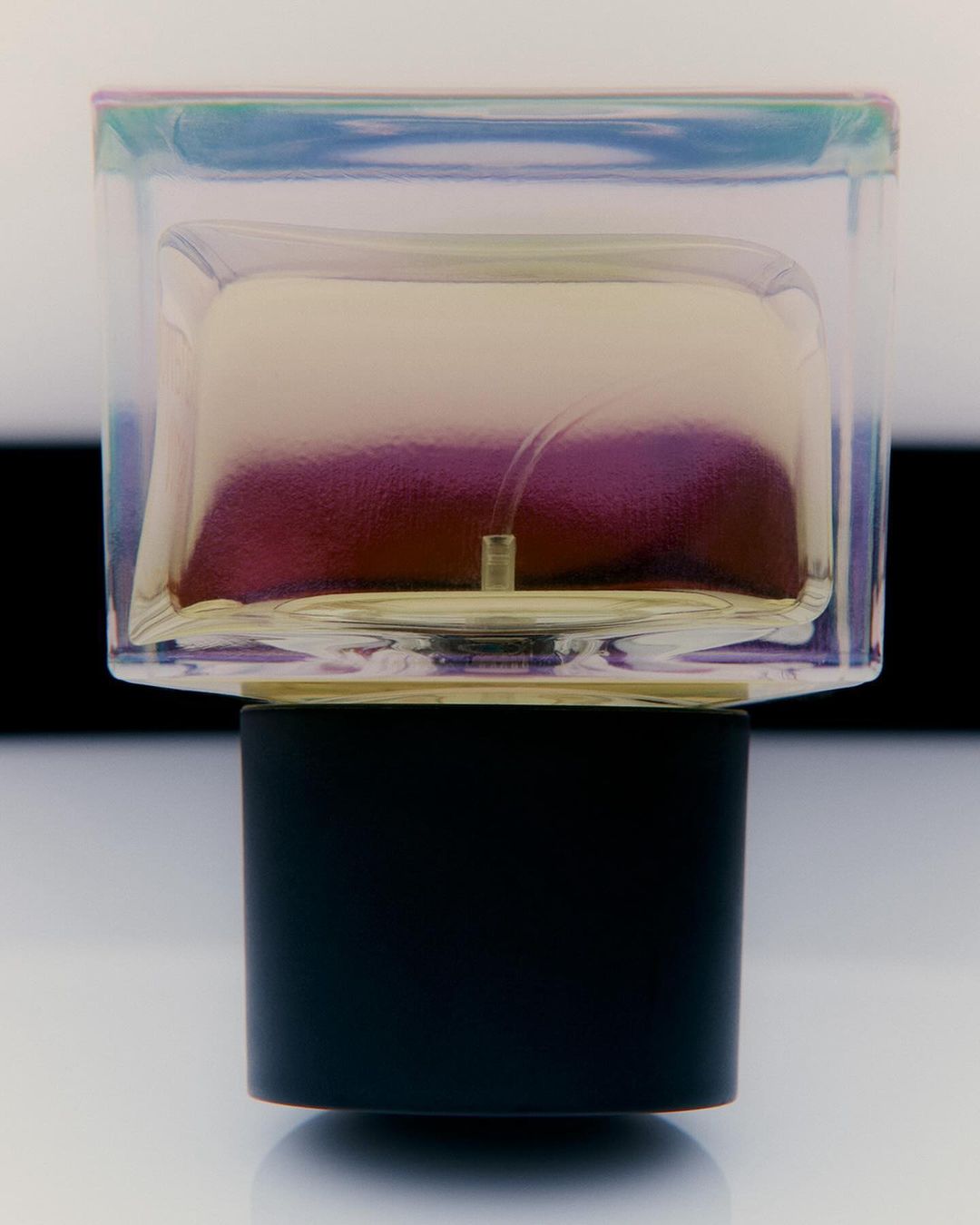 Close-up studio shot of a perfume bottle with a rectangular transparent glass body and a cylindrical black base on a reflective surface.