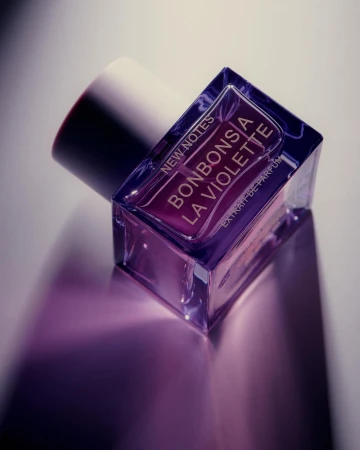 Close-up angled product shot of a square purple perfume bottle with white cap on a neutral gradient surface.