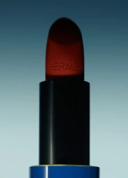 Close-up of a Hermes lipstick tube with embossed logo against a blue gradient background by Sara Zanoni