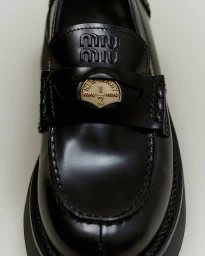 Close-up of a black patent leather loafer featuring a metallic coin detail and embossed branding on the strap, set against a neutral background.
