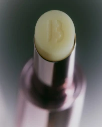 Close up product shot of a pale yellow lip balm stick in a metallic pink tube with an embossed letter B logo