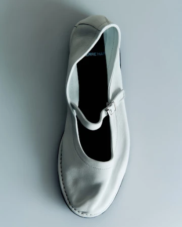 Top-down view of a white leather Mary Jane flat shoe with a buckled strap against a plain light grey studio background.