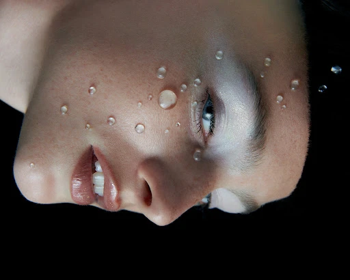 Close-up portrait of a woman with water droplets on her face against a black background by Sara Zanoni