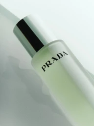 Close-up product shot of a frosted glass Prada fragrance bottle with a metallic cap on a pale surface