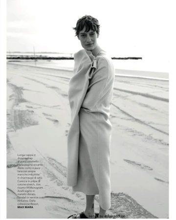 Model wrapped in a long coat standing on a sandy beach in a monochrome fashion editorial photograph by Stefano Galuzzi.