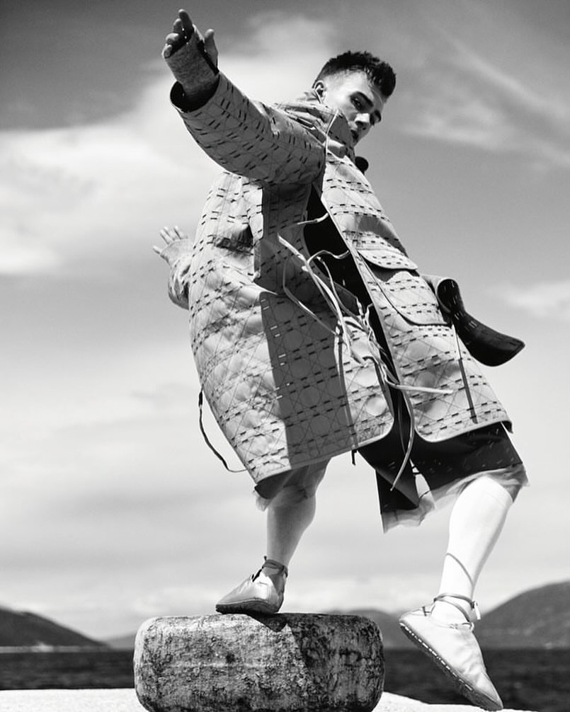 Monochrome portrait of a male fashion model wearing a quilted jacket and shorts, balancing on a round stone with a cloudy sky backdrop.