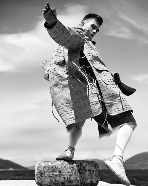 Monochrome portrait of a male fashion model wearing a quilted jacket and shorts, balancing on a round stone with a cloudy sky backdrop.