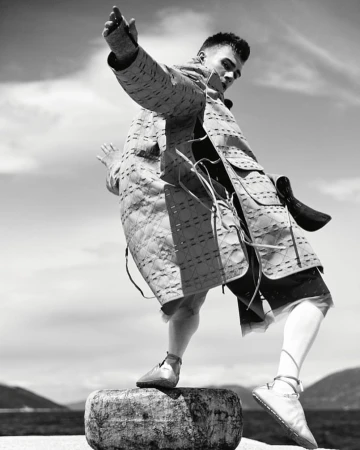 Monochrome portrait of a male fashion model wearing a quilted jacket and shorts, balancing on a round stone with a cloudy sky backdrop.