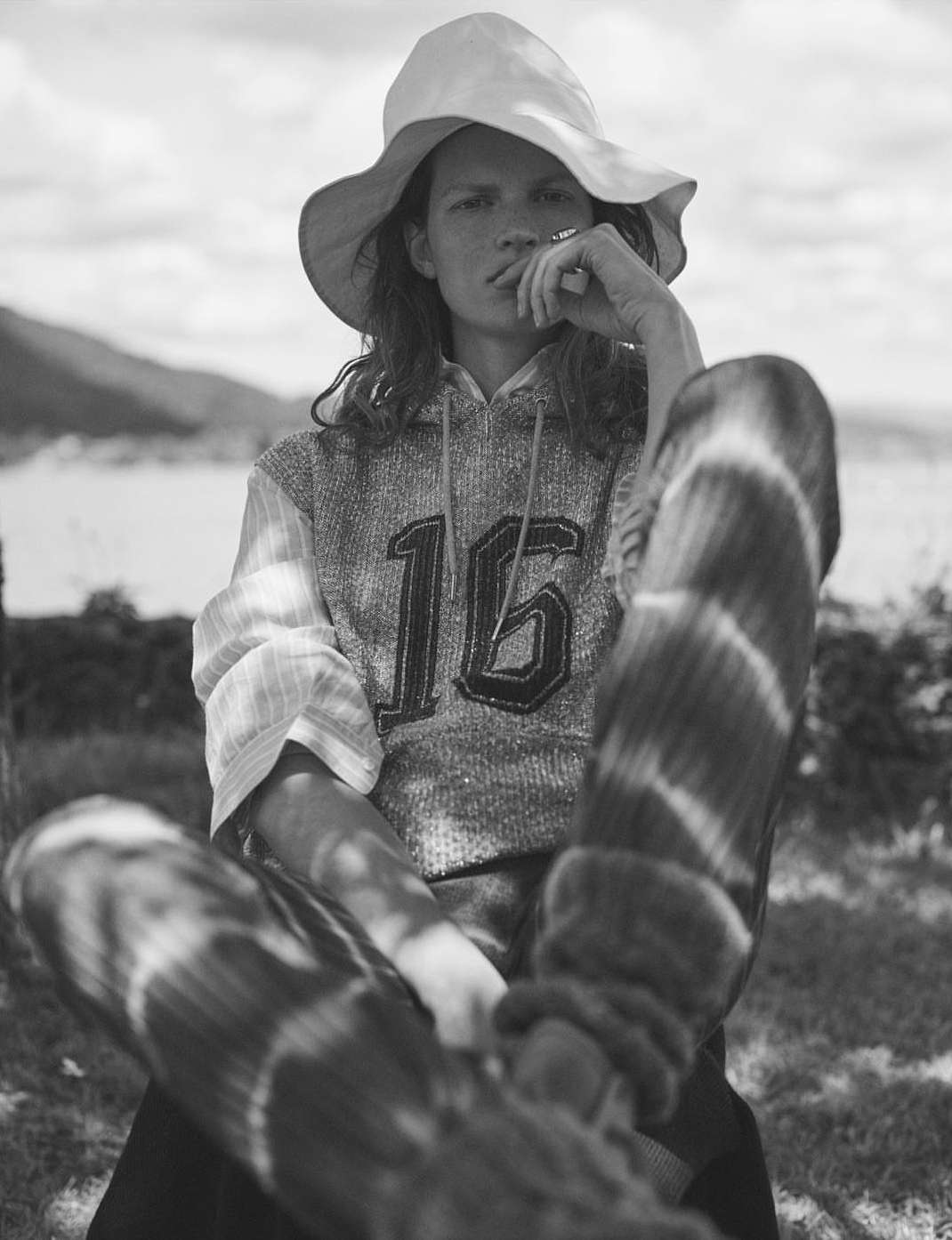 Black-and-white portrait of a young adult with a hat and hoodie with the number 16, sitting outdoors with a lake and trees in the background.