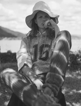 Black-and-white portrait of a young adult with a hat and hoodie with the number 16, sitting outdoors with a lake and trees in the background.