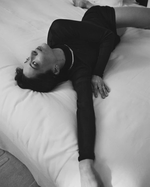 Black and white portrait of a woman reclining on a white bed, wearing a black dress and silver necklace, eyes looking upwards.