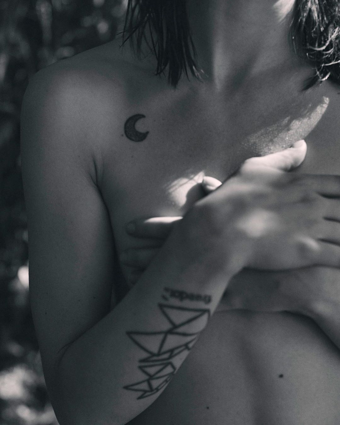 Black and white studio shot of a nude adult female torso with tattoos, posing in a natural outdoor setting with trees in the background.