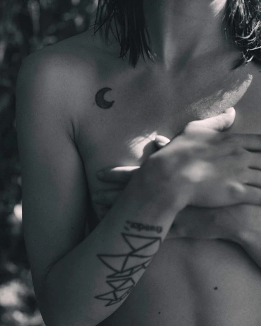 Black and white studio shot of a nude adult female torso with tattoos, posing in a natural outdoor setting with trees in the background.