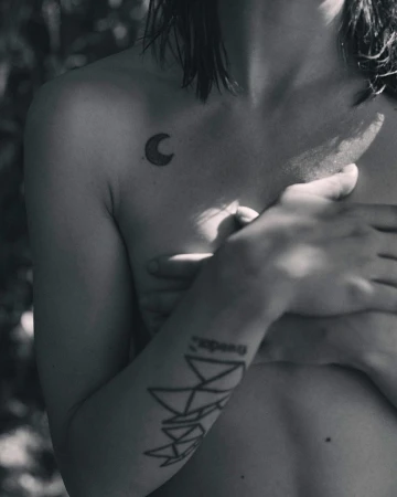 Black and white studio shot of a nude adult female torso with tattoos, posing in a natural outdoor setting with trees in the background.