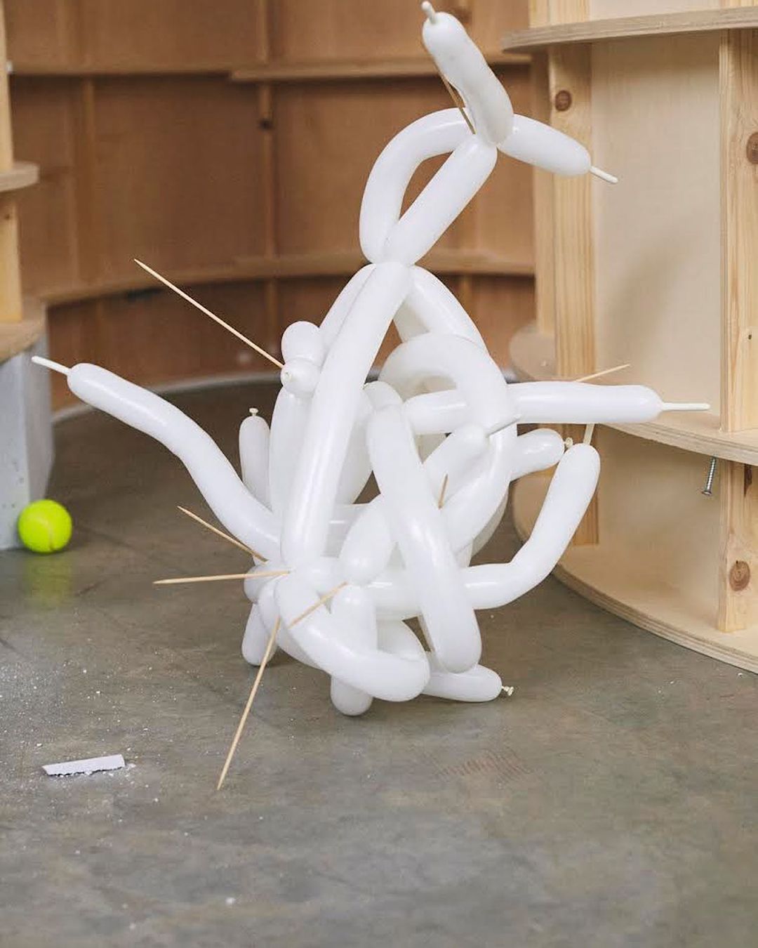 Abstract balloon sculpture on a concrete floor with toothpicks, against a background of light wood shelves and a yellow tennis ball.