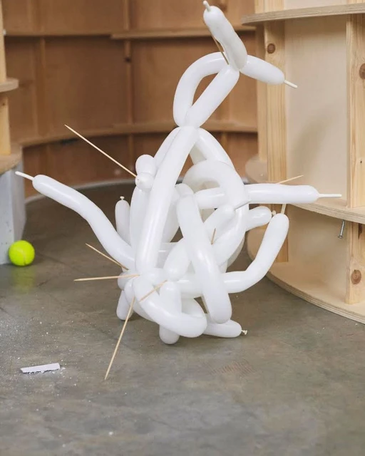Abstract balloon sculpture on a concrete floor with toothpicks, against a background of light wood shelves and a yellow tennis ball.