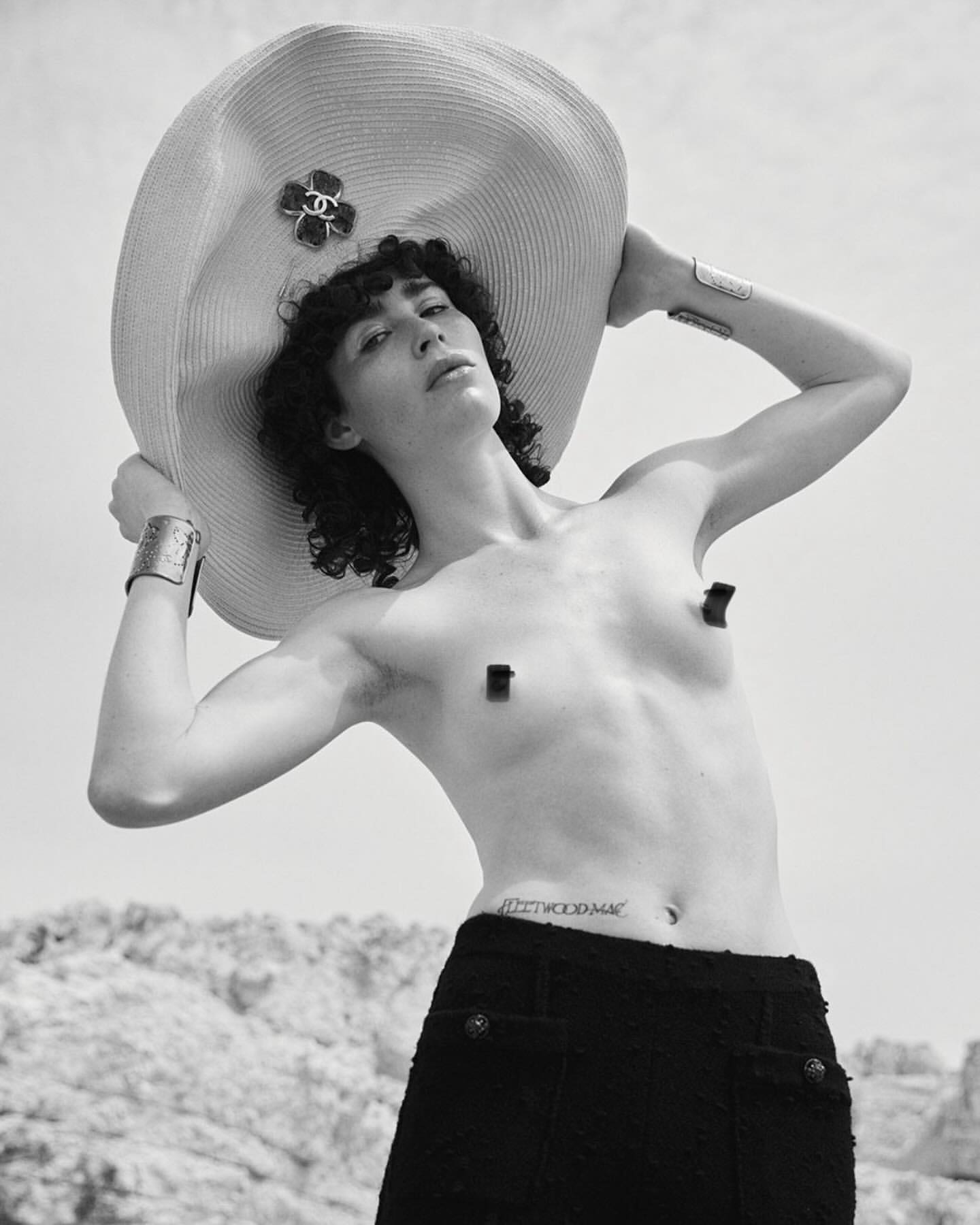 Person poses in a black skirt with a large straw hat on a bright, sunny day. The individual is standing outside near a rocky hill.